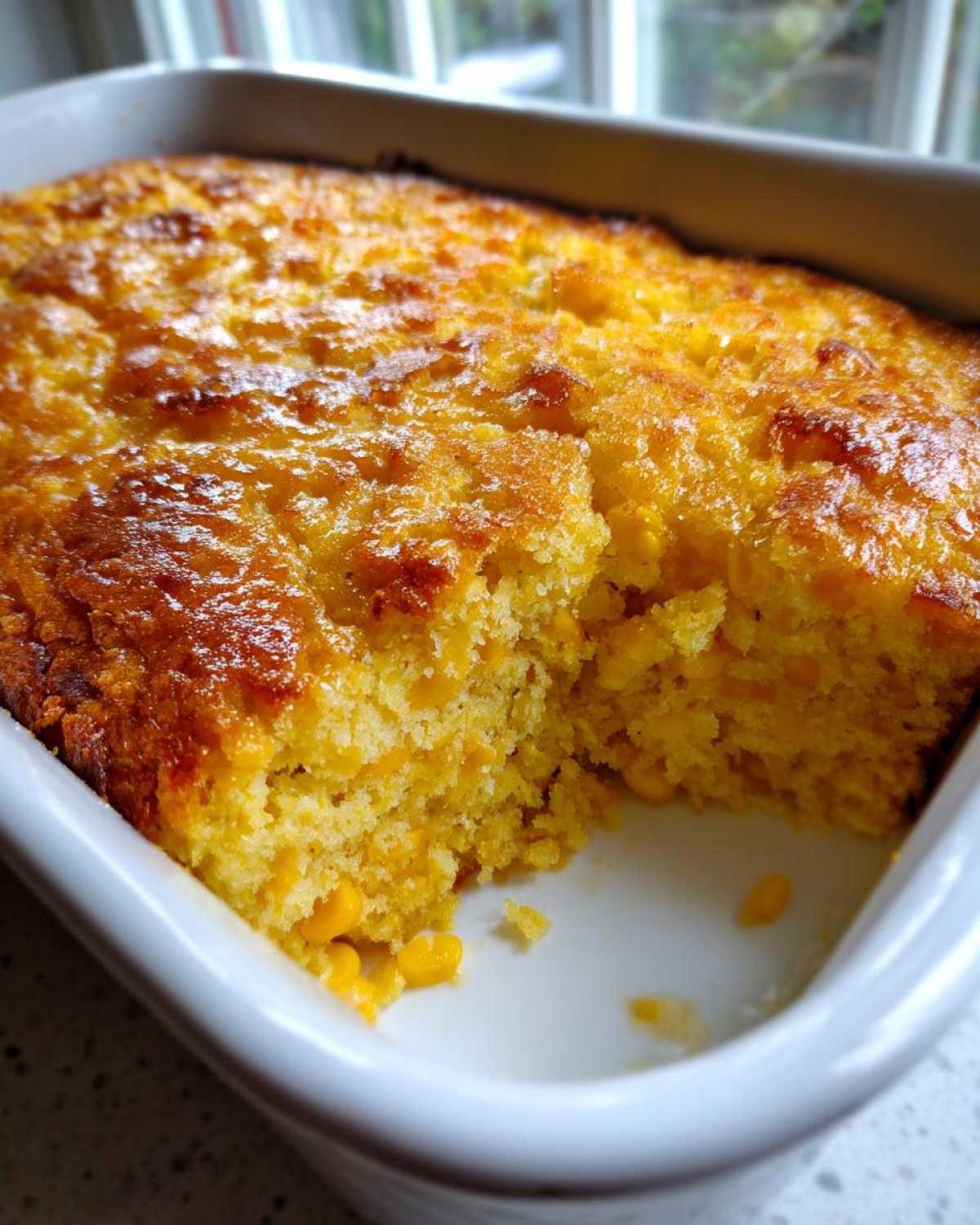 A golden-brown, moist slice taken out of a freshly baked jiffy corn casserole in a white baking dish.