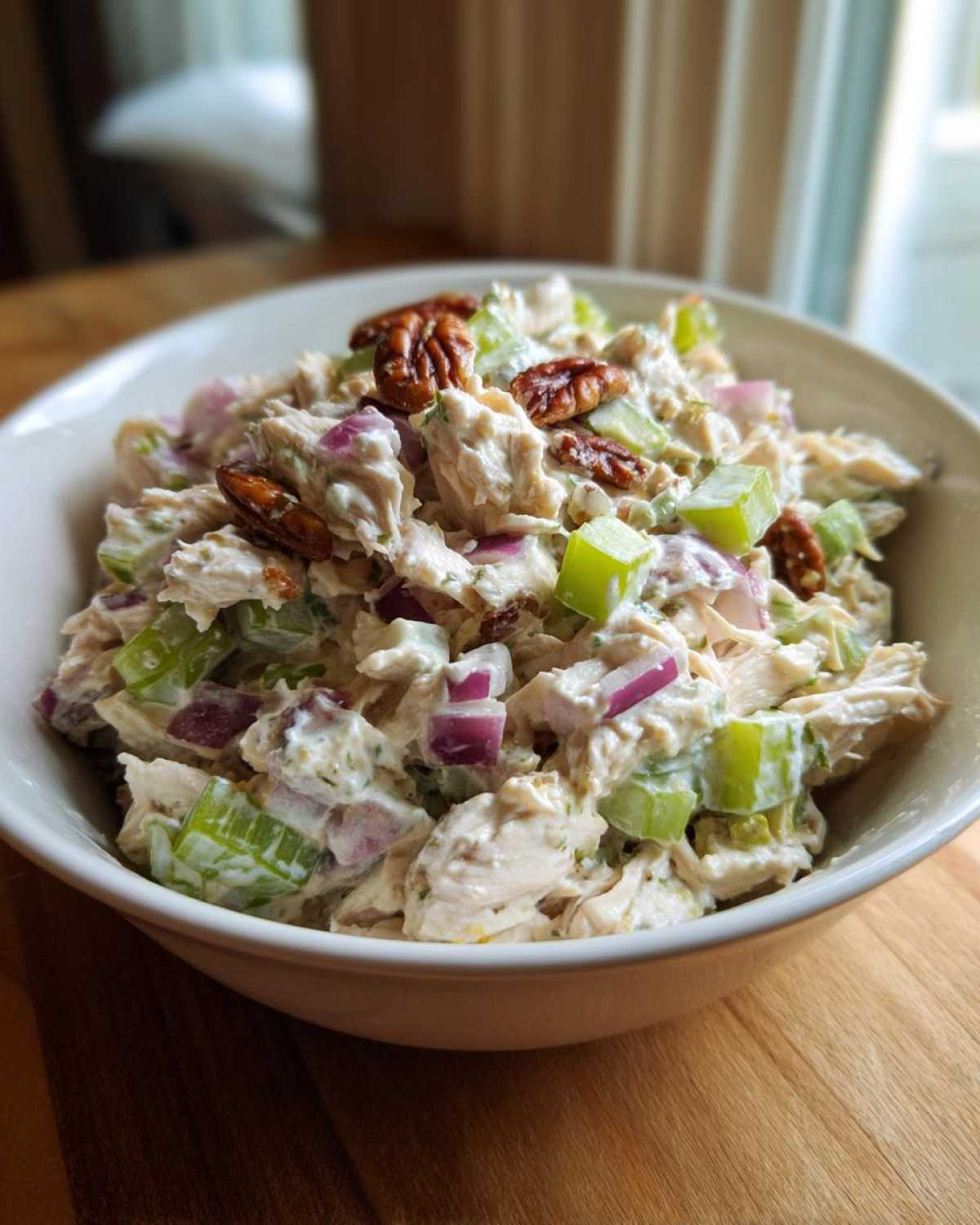 A close-up of creamy Leftover Rotisserie Chicken Salad mixed with celery, red onion, and topped with pecans in a white bowl.
