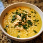A close-up of a white bowl filled with rich, yellow Mediterranean Lemon Chicken Soup, featuring shredded chicken, rice, and fresh parsley garnish.