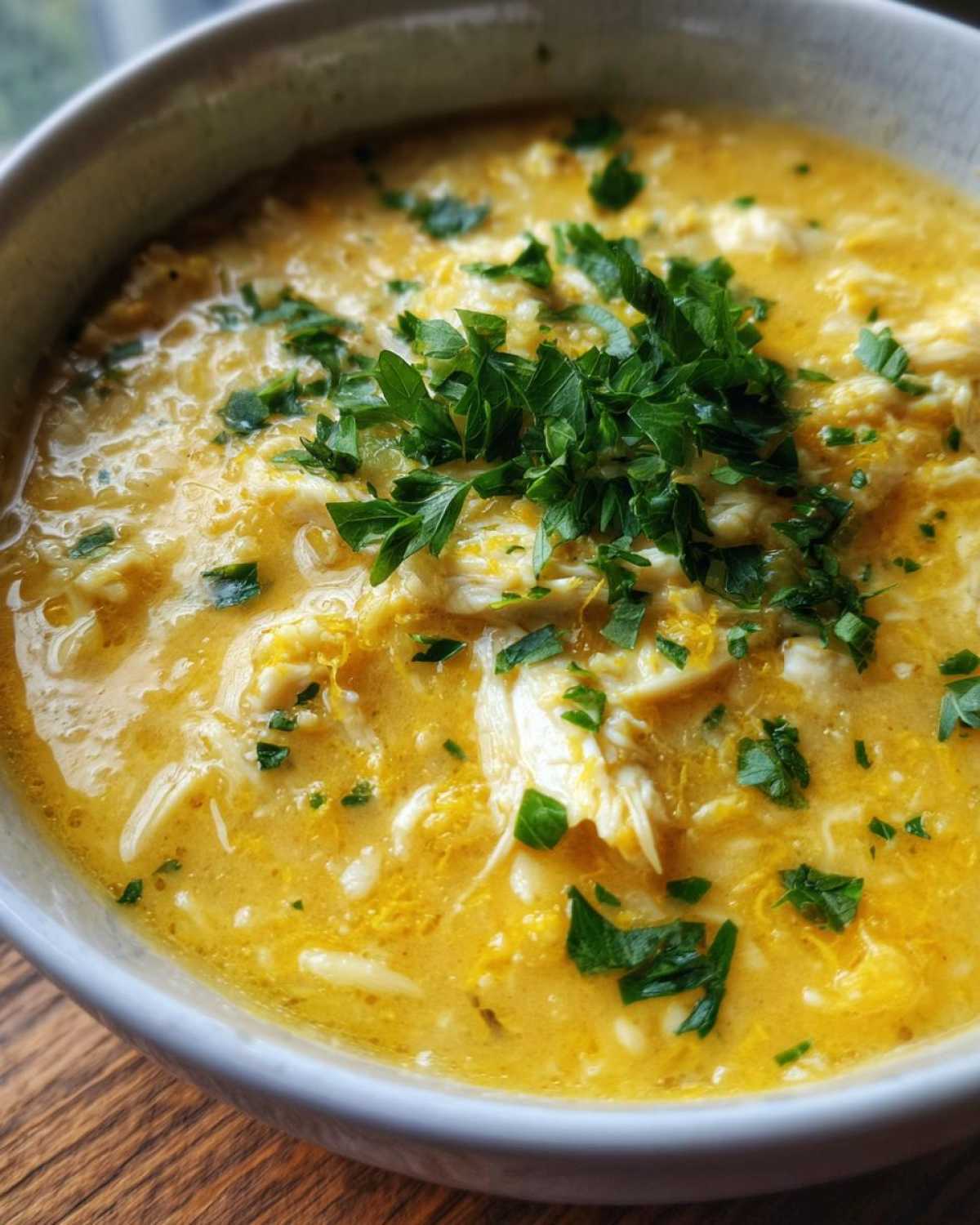 Close-up of a bowl of creamy, yellow Mediterranean Lemon Chicken Soup topped with shredded chicken and fresh parsley.