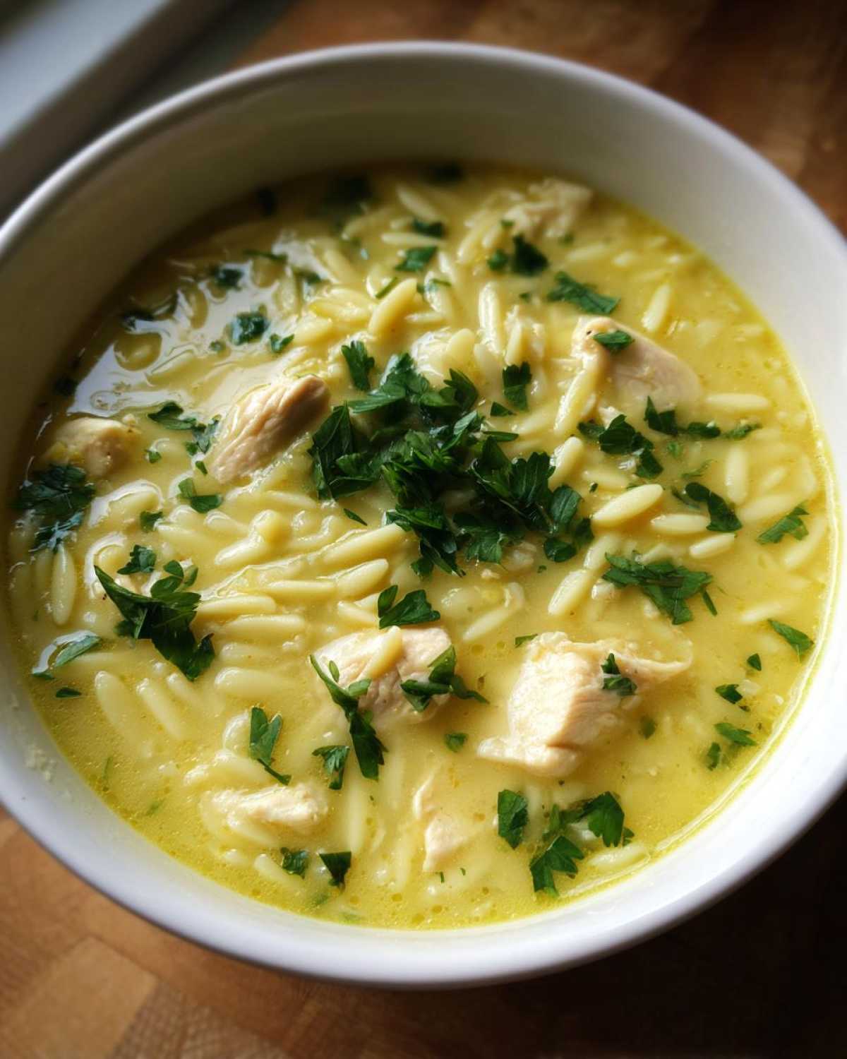 Close-up of a white bowl filled with vibrant Mediterranean Lemon Chicken Soup, featuring orzo pasta, chicken pieces, and fresh parsley.