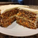 A close-up of a slice of moist carrot cake showing shredded carrots, nuts, and thick cream cheese frosting.