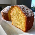 Close-up of a bright yellow slice of olive oil cake dusted with powdered sugar on a white plate.