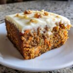 A close-up of a moist slice of carrot cake topped with thick cream cheese frosting and chopped walnuts.