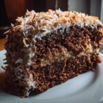 Close-up of a rich slice of german chocolate cake layered with coconut-pecan filling and topped with toasted coconut flakes.