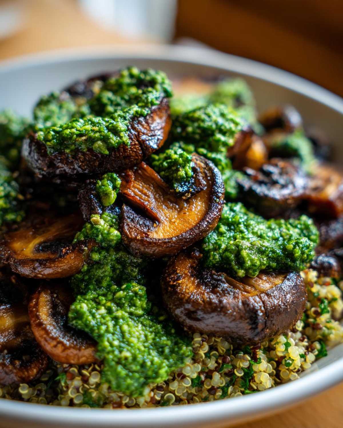 Close-up of savory, seared mushrooms topped with bright green kale pesto, served over a bed of tri-color quinoa in a white bowl.