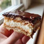 A person holding a layered slice of no-bake eclair cake featuring graham crackers, creamy filling, and a rich chocolate topping.