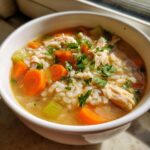A close-up of a white bowl filled with Nourishing Chicken and Rice Soup, featuring shredded chicken, rice, carrots, and celery, topped with parsley.