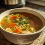 Close-up of a white mug filled with Nourishing Chicken and Rice Soup, featuring shredded chicken, bright orange carrots, celery, and parsley garnish.