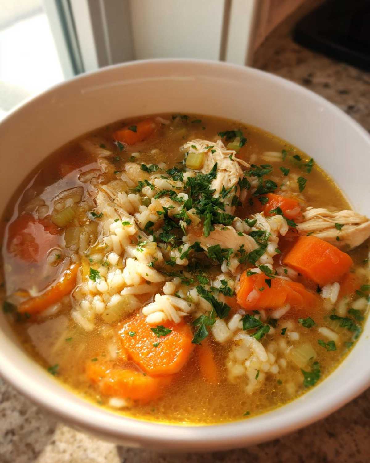 A close-up of a white bowl filled with Nourishing Chicken and Rice Soup, featuring shredded chicken, rice, carrots, and parsley.