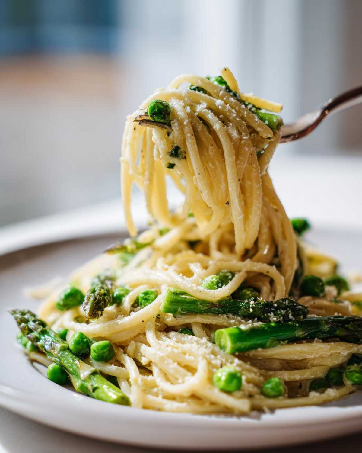 A fork lifting spaghetti from a plate of One Pot Spring Pasta Primavera, featuring bright green peas and asparagus spears, topped with Parmesan.