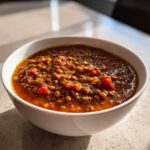 A close-up of a rich, thick serving of Pantry Staples Lentil Stew in a white bowl, highlighted by natural sunlight.