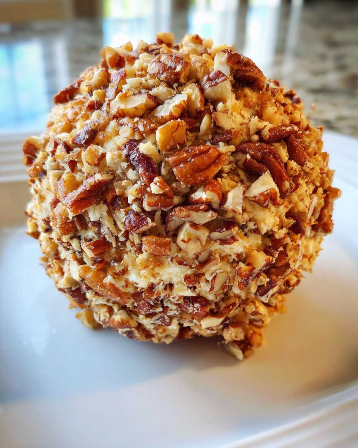 A close-up of a perfectly formed cheese ball recipe coated entirely in chopped pecans, resting on a white plate.