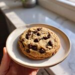 A perfectly baked, thick Chocolate Chip Cookie held on a small ceramic plate in bright sunlight.