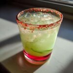 A chilled Pickle Margarita cocktail in a glass with a bright red chili salt rim, filled with ice.