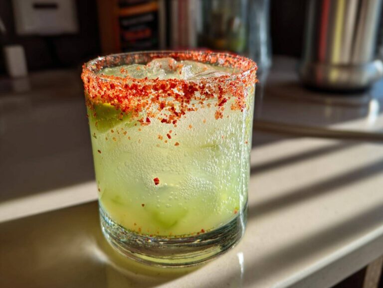 Close-up of a chilled Pickle Margarita with a vibrant red chili-salt rim, sitting on a sunlit countertop.