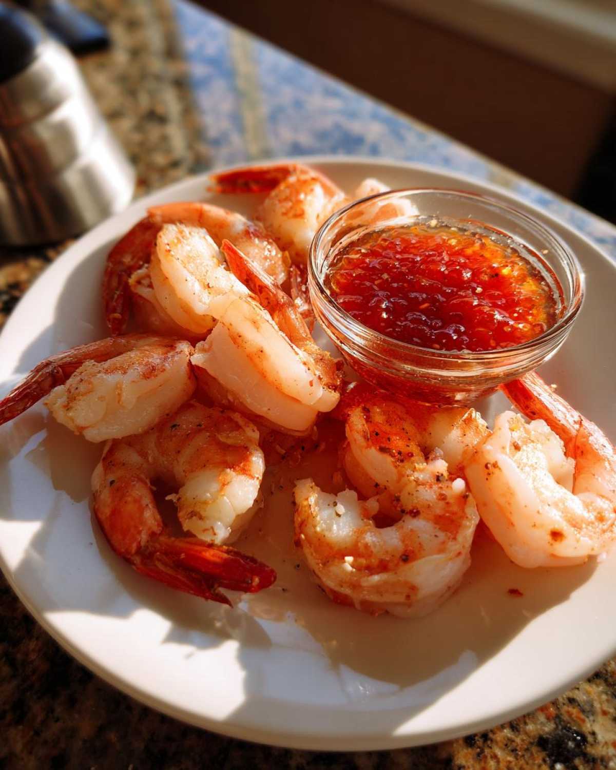 A white plate holding several cooked shrimp surrounding a small glass bowl of bright red, chunky shrimp cocktail sauce.
