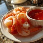 Close-up of cooked, peeled shrimp served on a white plate next to a ramekin of red dipping sauce for shrimp cocktail.