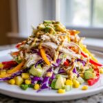 A colorful mound of Rainbow Chicken Salad Honey Mustard featuring shredded chicken, purple cabbage, corn, peppers, and avocado, drizzled with dressing.