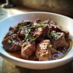Close-up of tender chunks of Roman Lamb Stew (Abbacchio) garnished with fresh rosemary sprigs in a white bowl.