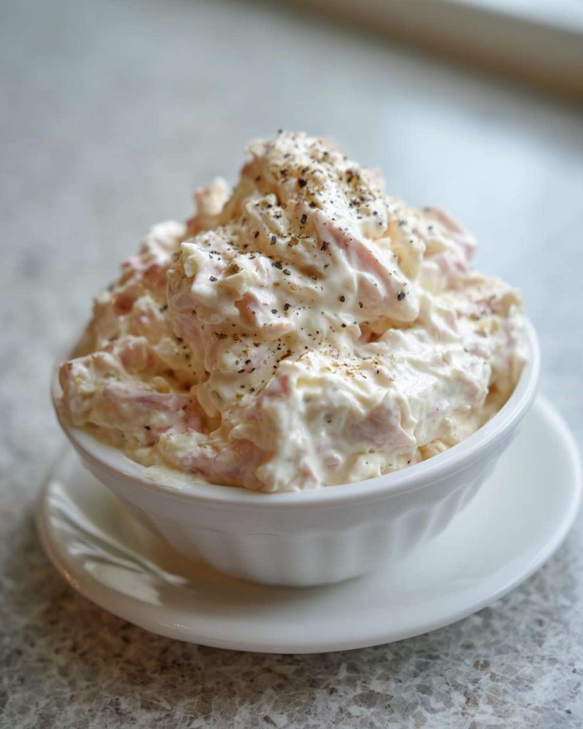 A white bowl filled high with creamy ham salad recipe, generously topped with cracked black pepper.