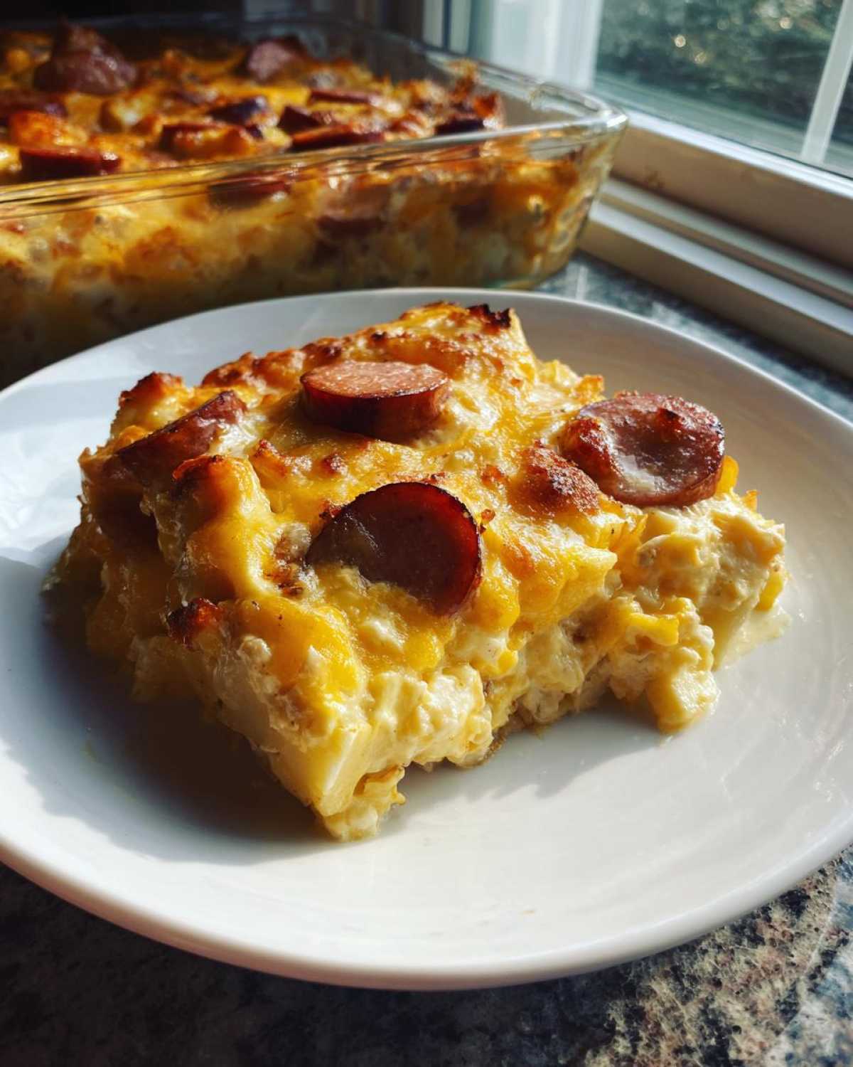 A hearty slice of pierogi casserole topped with melted cheddar cheese and sliced smoked sausage on a white plate.