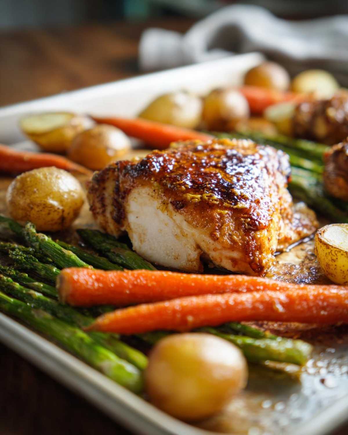 Close-up of a perfectly cooked piece of Sheet Pan Easter Dinner Chicken surrounded by roasted asparagus, carrots, and baby potatoes.