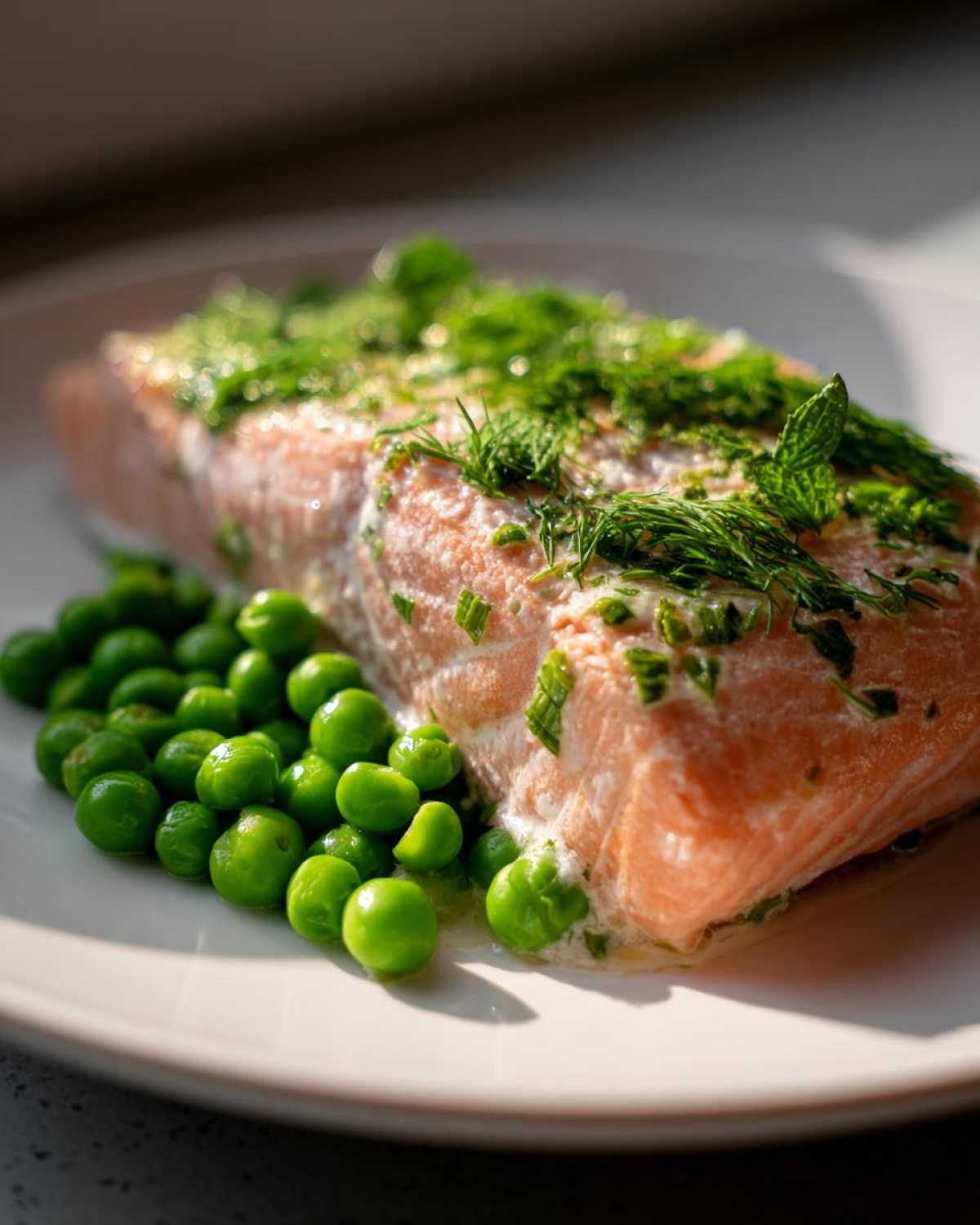 Close-up of a cooked salmon fillet topped with fresh dill and mint, served next to bright green spring peas.