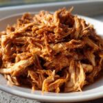 Close-up of a mound of tender, shredded bbq chicken coated in rich sauce, served on a white plate.