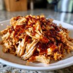 Close-up of shredded bbq chicken generously topped with rich, dark barbecue sauce on a white plate.