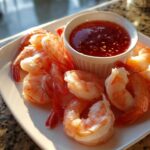 A plate of perfectly cooked shrimp cocktail served with a small white ramekin of sweet chili dipping sauce.