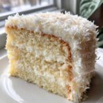 Close-up of a moist slice of coconut cake recipe, layered with white frosting and topped generously with shredded coconut.