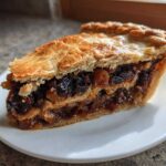 A close-up of a thick slice of mincemeat pie showing the dark, rich fruit filling and golden, flaky pastry crust.