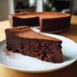 A thick slice of rich flourless chocolate cake dusted with cocoa powder on a white plate.