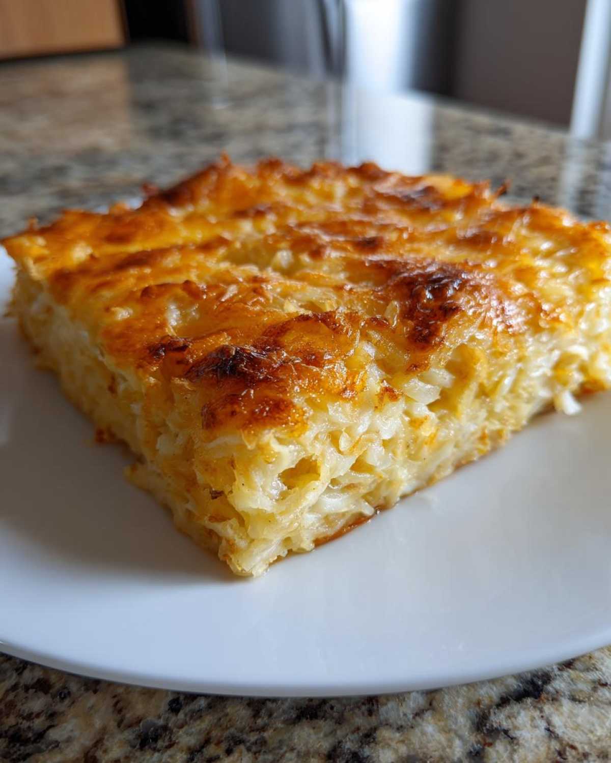A close-up of a square slice of cheesy, golden-brown hashbrown casserole on a white plate.