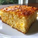 A close-up of a moist, square slice of golden jiffy corn casserole with visible corn kernels, sitting on a white plate.
