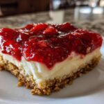 A close-up of a square slice of pretzel salad showing the bottom pretzel crust, creamy layer, and bright red strawberry topping.