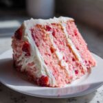 A close-up of a moist slice of pink strawberry cake layered with white frosting and visible strawberry pieces.