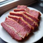 Close-up of several thick, juicy slices of pink corned beef with a fat cap, arranged on a white plate.