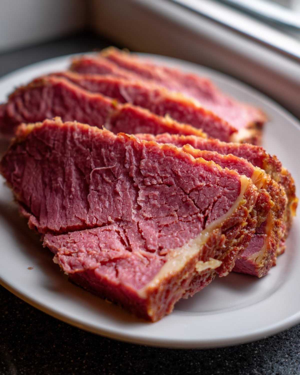 Close-up of several thick, tender slices of pink corned beef resting on a white plate.