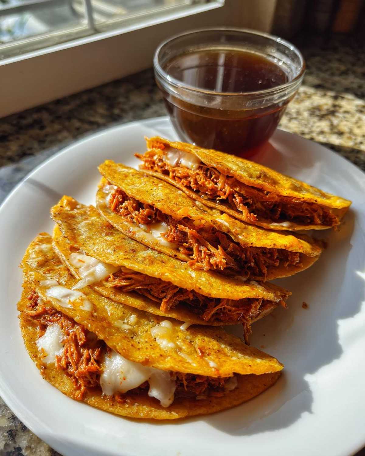 Four crispy, cheesy Slow Cooked Birria Tacos served on a white plate next to a small bowl of dark consommé.