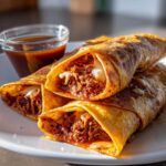 Three golden, crispy Slow Cooked Birria Tacos filled with shredded meat and cheese, served with a side of dark consommé.