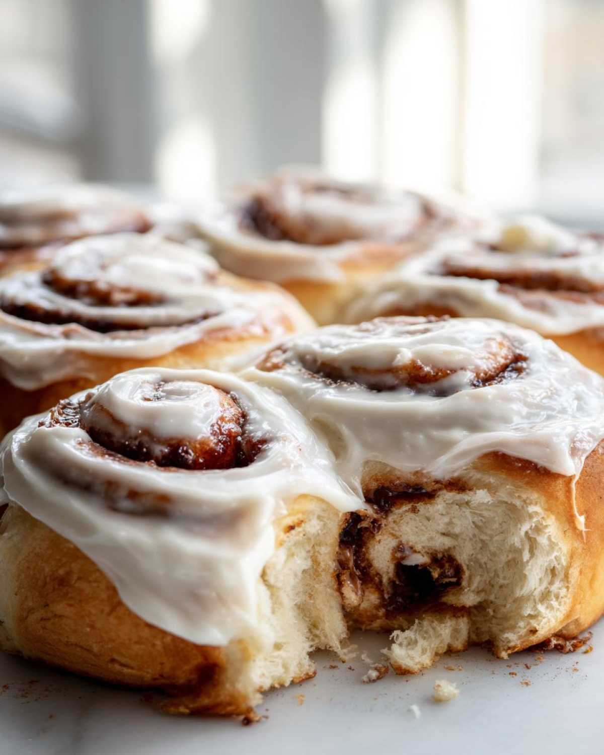 A close-up of several soft, freshly baked cinnamon buns topped with thick white icing, one bun pulled apart to show the fluffy interior.