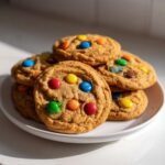 Close-up of several soft m&m cookies stacked on a white plate, featuring colorful candies.