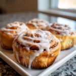 Close-up of four freshly baked Sourdough Cardamom Buns topped with thick white icing, sitting on a white rectangular tray.