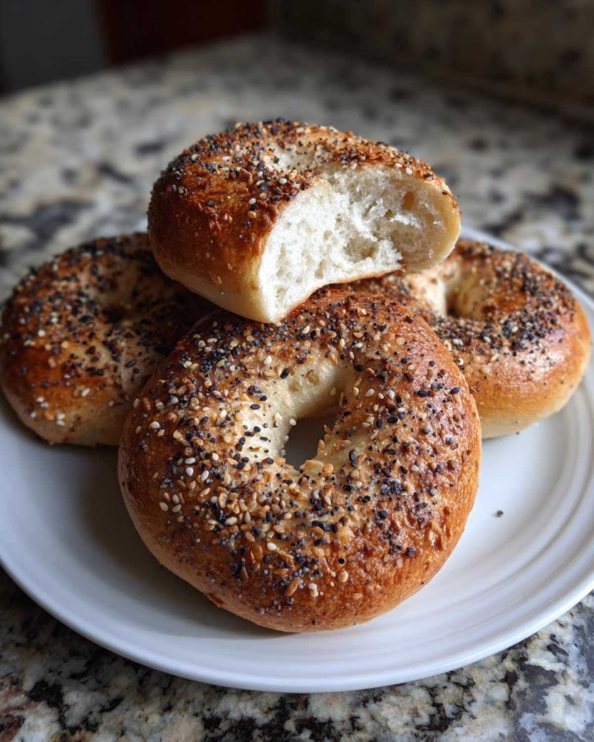 A stack of four everything sourdough discard bagels on a white plate, one cut open showing the fluffy interior.