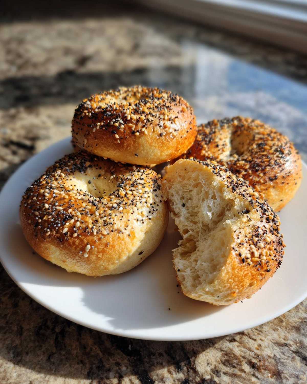 Three golden brown sourdough discard bagels on a white plate, one is broken open showing the chewy interior.