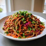 A close-up of a mound of Spicy Peanut Soba Noodle Salad tossed with shredded carrots and red cabbage, topped with green onions.