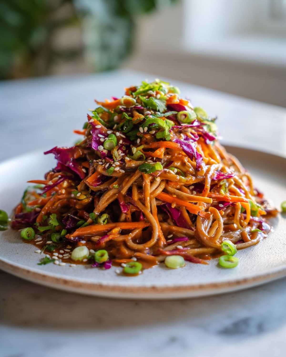 A mound of Spicy Peanut Soba Noodle Salad tossed in peanut sauce, topped with shredded cabbage, carrots, cilantro, and sesame seeds.