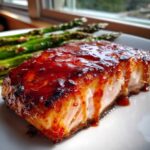 Close-up of a perfectly cooked Spicy Salmon fillet covered in a thick, glossy red glaze, served with grilled asparagus.
